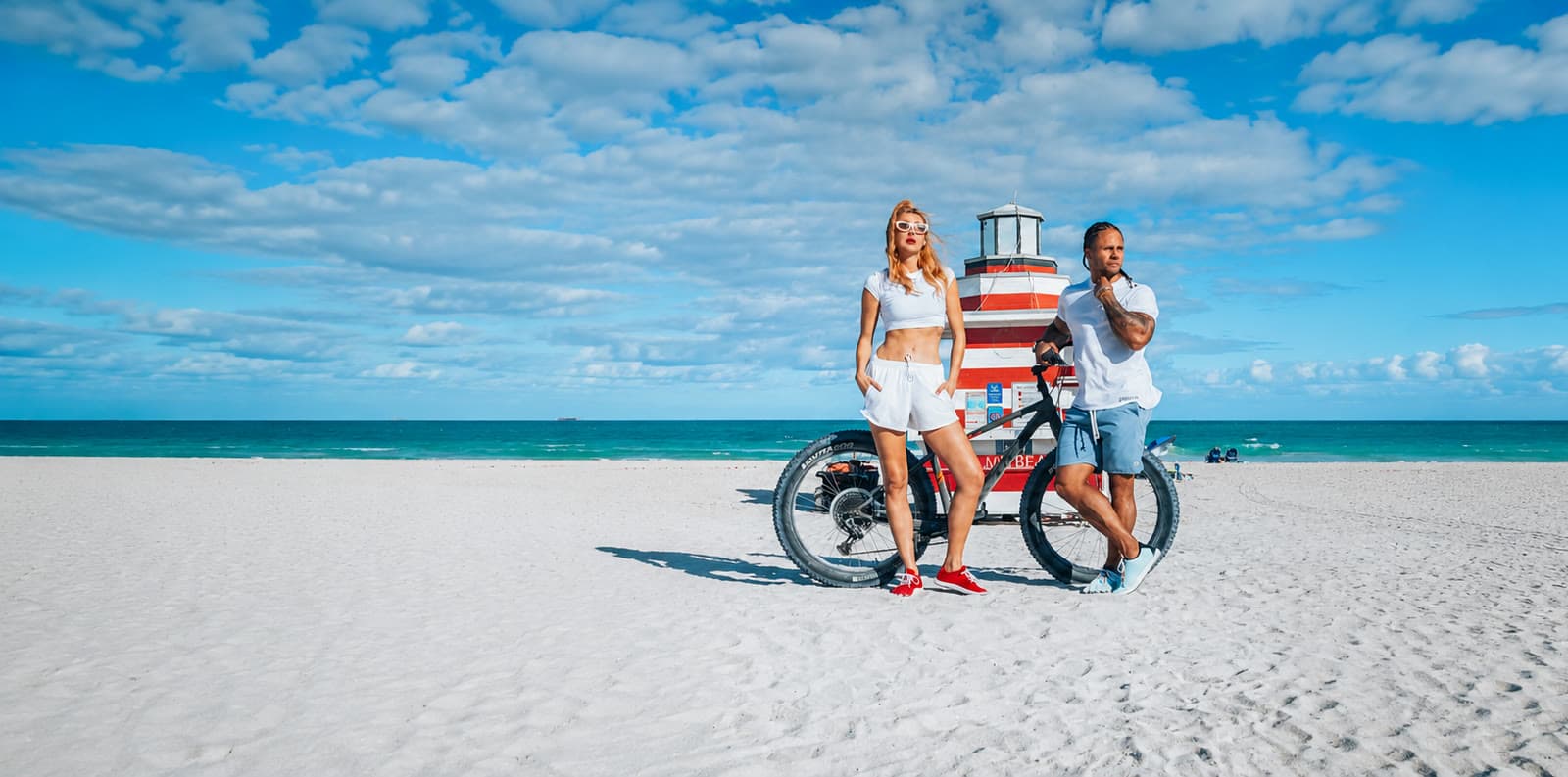 A man and woman on Miami Beach beside a red-and-white striped lighthouse, wearing Peluva barefoot shoes in red and light blue